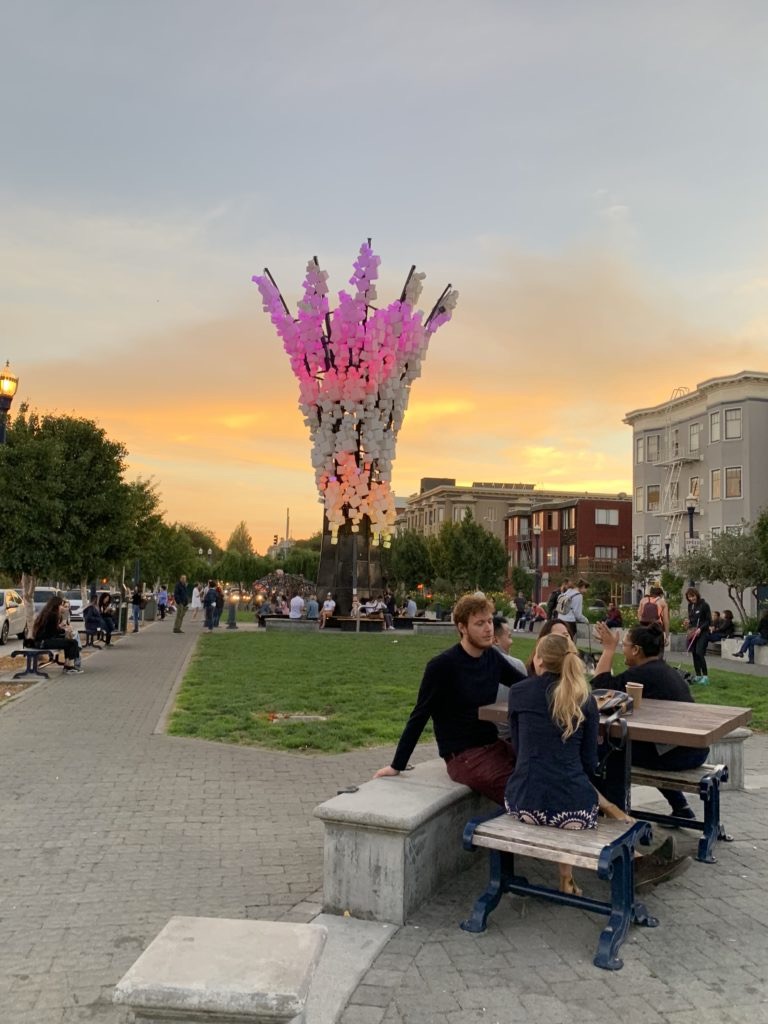 Piazza con al centro una installazione colorata e ragazzi sulle panchine intorno al tramonto