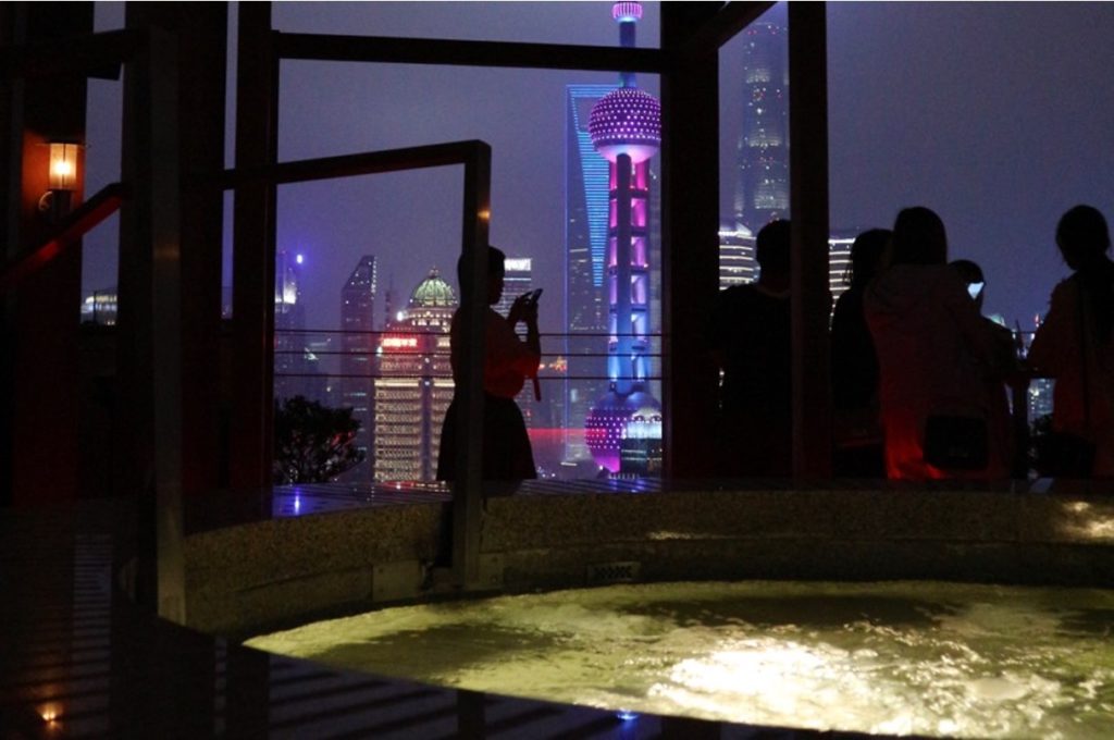 Primo piano di una Jacuzzi illuminata di notte, su una terrazza con vista sui grattacieli di Shanghai, con sagome nere di persone