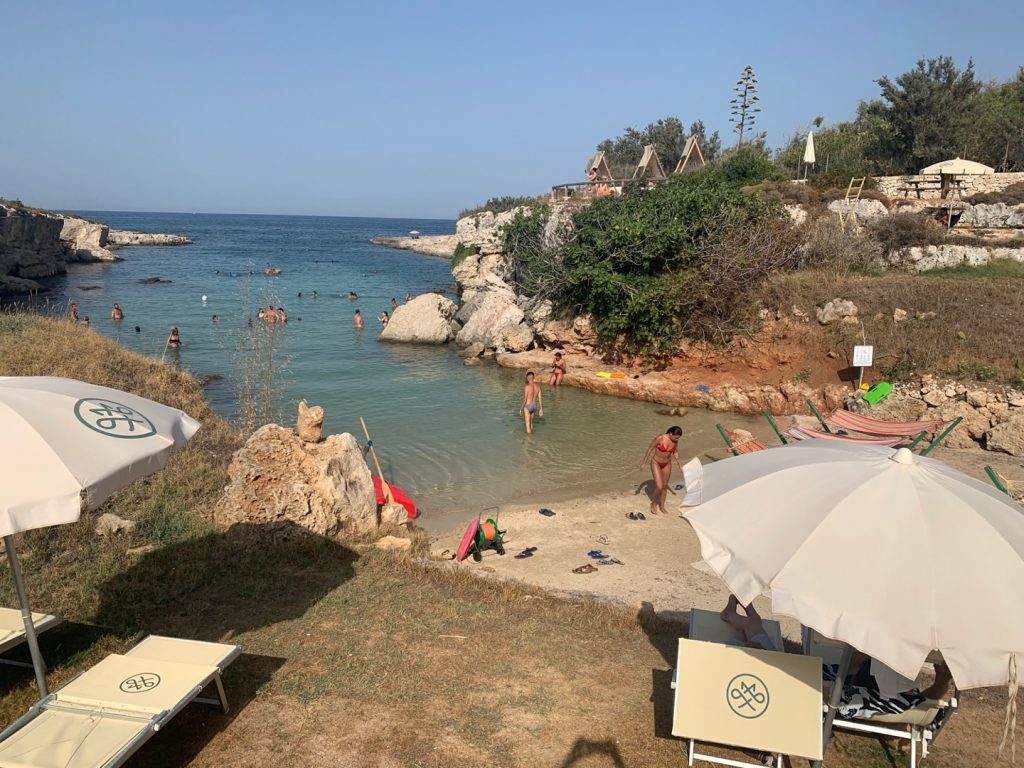 Caletta con persone che fanno il bagno nel mare e ombrelloni e lettini sull'erba vicino alla spiaggia
